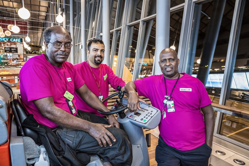 Tre män i rosa tröjor vid en städmaskin