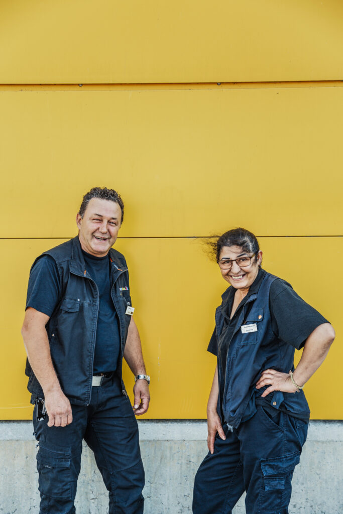 En man och kvinna i Samhallkläder ler mot kameran framför en gul husvägg