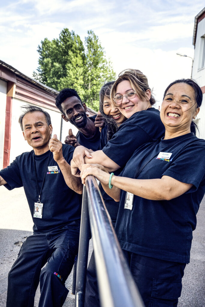 Ett gäng Samhall-arbetare står vid ett räcke och hänger och ler inför kameran