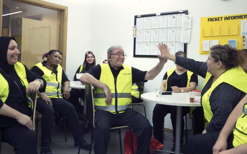 Samhallarbetare sitter i grupprum. Två av dem ger high five till varandra