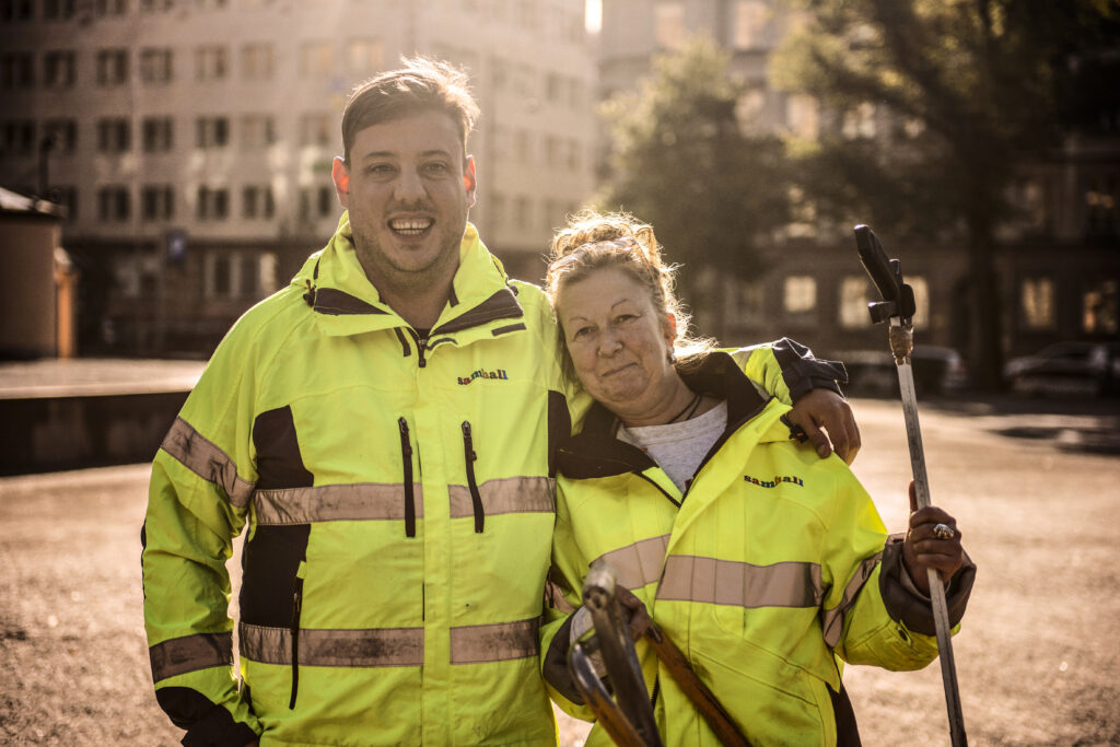 Två Samhall medarbetare i reflex arbetskläder som står i motljus och ler in i kameran.