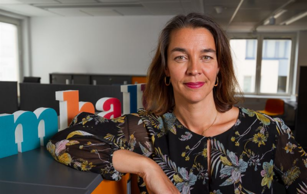 En kvinna med brunt hår lutar sig mot ett skåp och kollar in i kameran