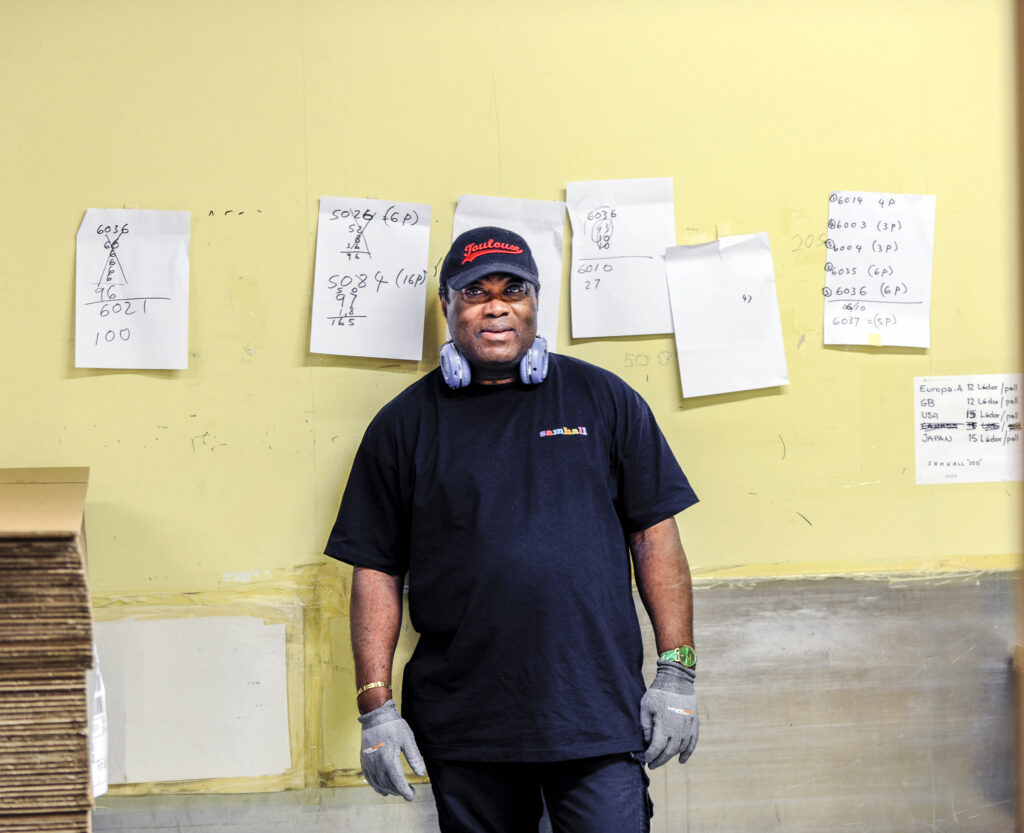 Person i arbetskläder och hörselskydd runt halsen posar framför en gul vägg med papper med uträkningar upsatta