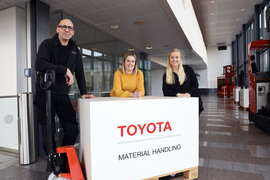 Bild på tre personer som står lutade på en kartong med loggan Toyota. Lådan står på en maskin som lyfter tunga objekt. 