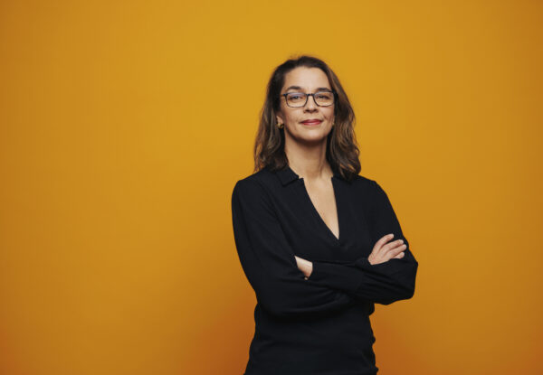 Porträtt av Sara Revell Ford framför en orange bakgrund. Hon bär svart kavaj och har armarna i kors.