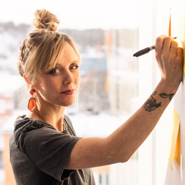 Kvinna tittar in i kameran och skriver på en whiteboard-tavla.