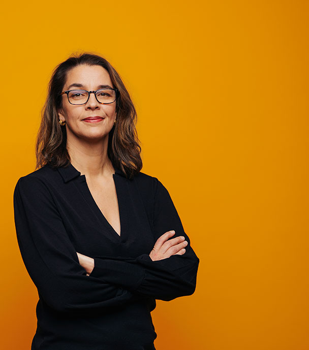 Portätt av Sara som står mot en orange bakgrund. Hon har armarna i kors och ser in i kameran.