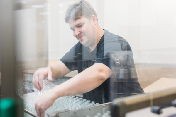 Andreas Gräntz matar flaskor i en maskin. Fotot är taget genom en glasruta.