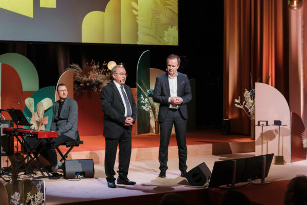 bild på tre män som står på scenen. Två står upp och dens tredje sitter ner bakom en keyboard. 