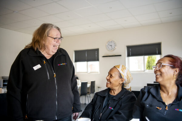 Annika pratar med två kollegor som sitter ner. De sitter i ett ljust rum med svarta persienner. Alla bär svarta arbetskläder. 