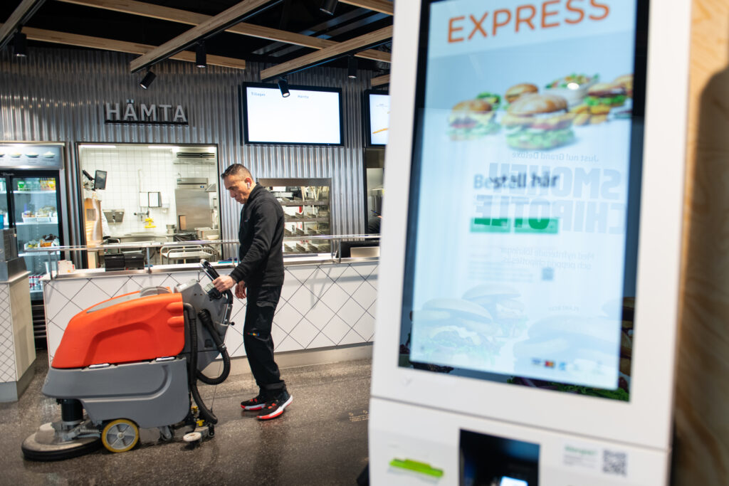 Humberto Rengifo går med en sopmaskin och tvättar golvet i en max-restaurang. I förgrunden syns en beställnignsmaskin. 