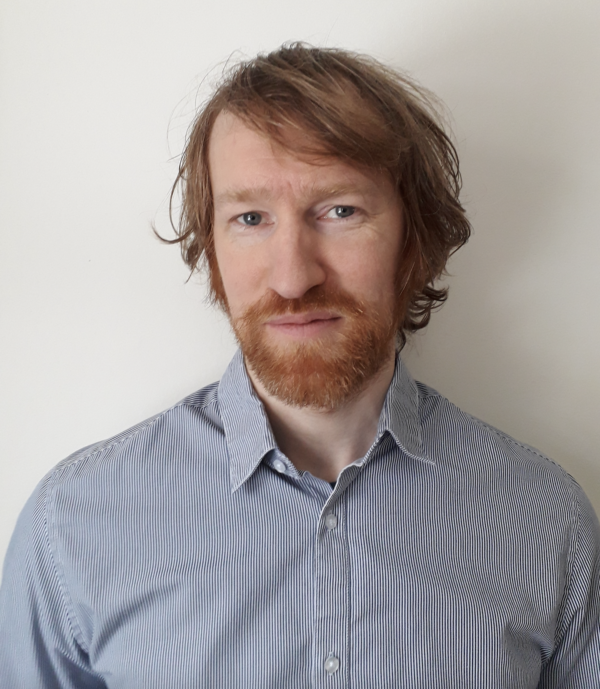 Johan Stenmark tittar in i kameran och bär en blå skjorta. Bakgrunden är vit och hans hår och skägg är brunt.