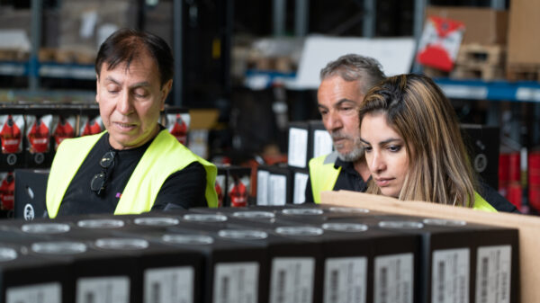 Två samhall-medarbetare och Nooshi står bakom några produkter. Man ser deras huvuden sticka upp bakom lådorna som täcker deras kroppar. De bär reflexvästar. 