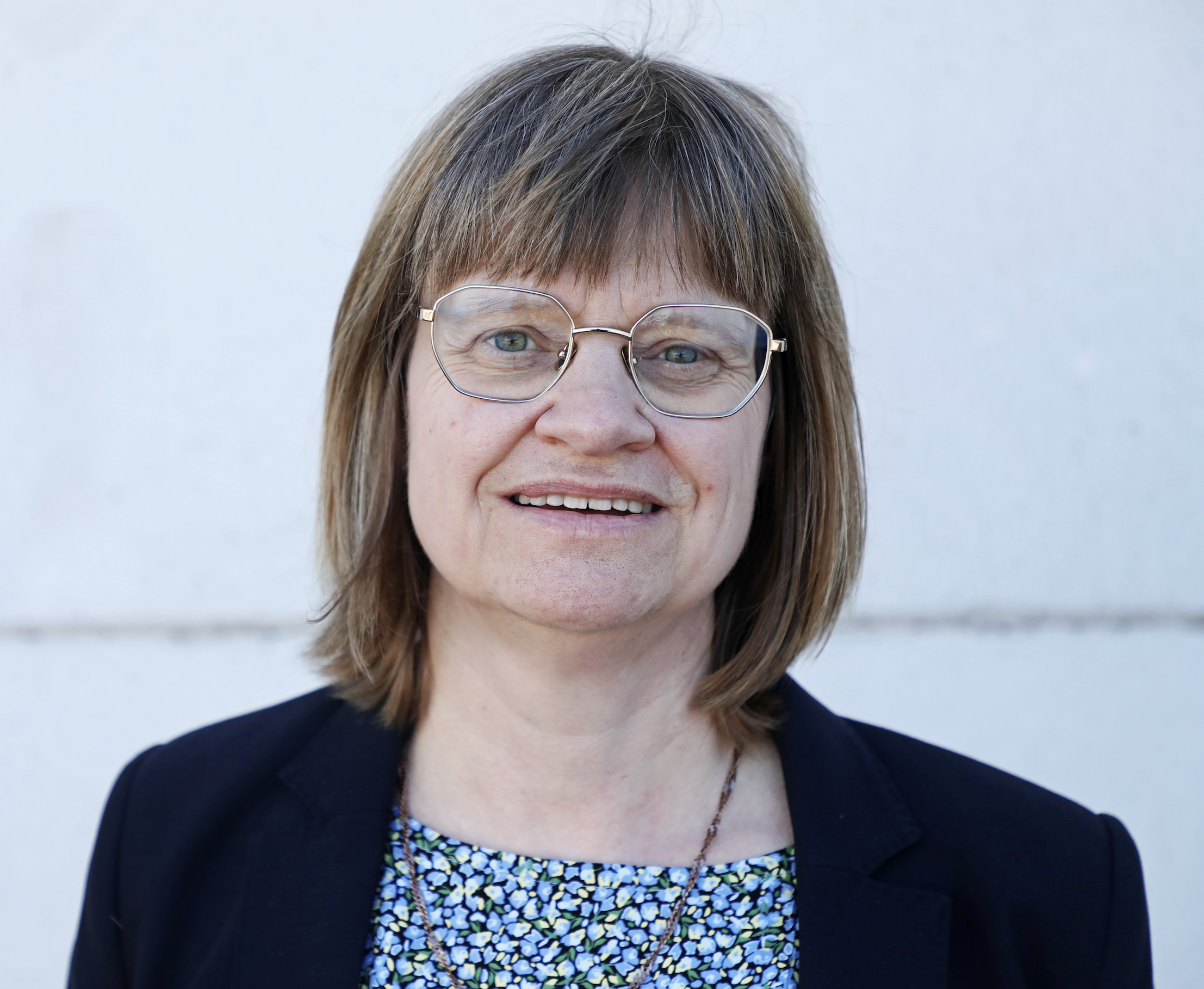 Porträtt på Karin mot en ljus fasadbakgrund utomhus.Hon bär glasögon och har svart kavaj.