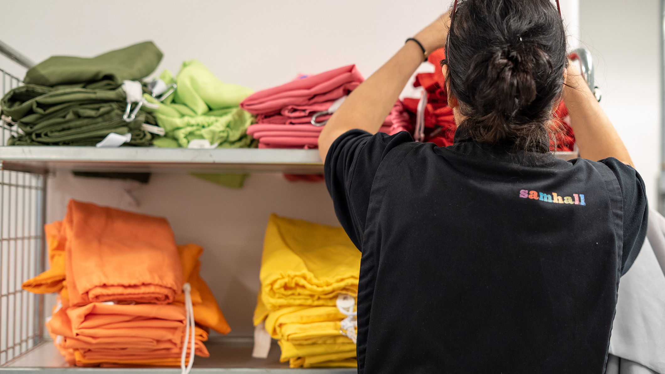 Person med mörkt hår uppsatt i en knut syns bakifrån. Hon har svart väst med Samhalls logga och står och viker färgglada tyger.