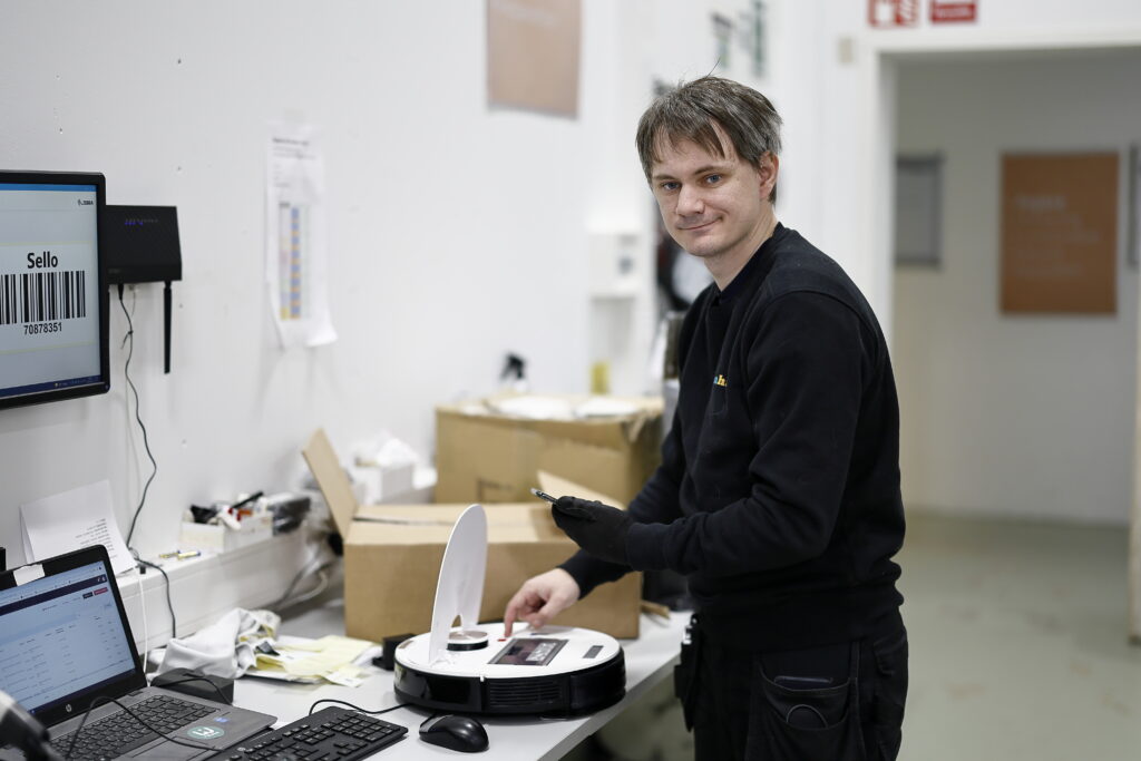 Medarbetaren Alexander står vid ett bord och mekar med en robotdammsugare. Han tittar in i kameran. 