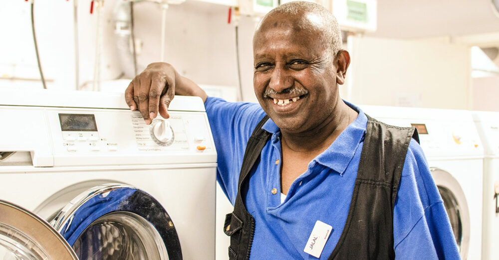 En person med rakat hår vid namn Jalal står lutad mot en tvättmaskin. Han har blå skjorta och svart väst samt namnbricka på bröstet. Han ler stort mot kameran.