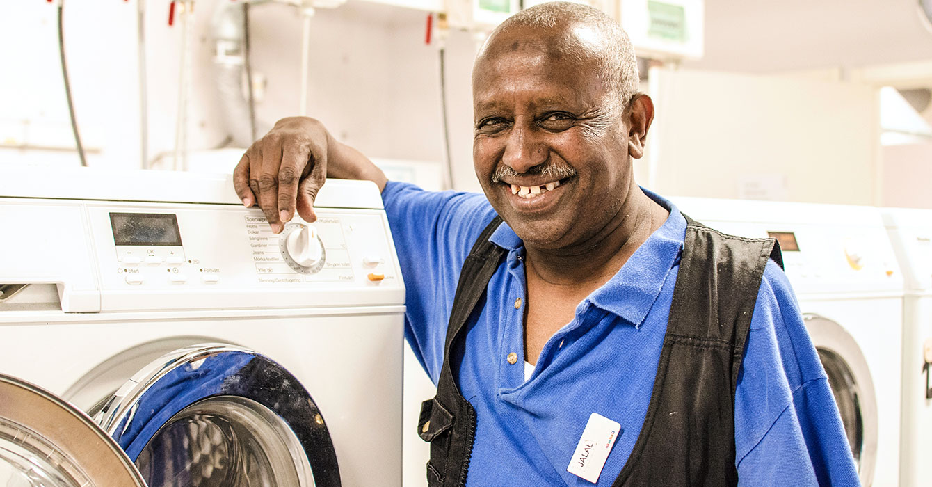En person med rakat hår vid namn Jalal står lutad mot en tvättmaskin. Han har blå skjorta och svart väst samt namnbricka på bröstet. Han ler stort mot kameran.