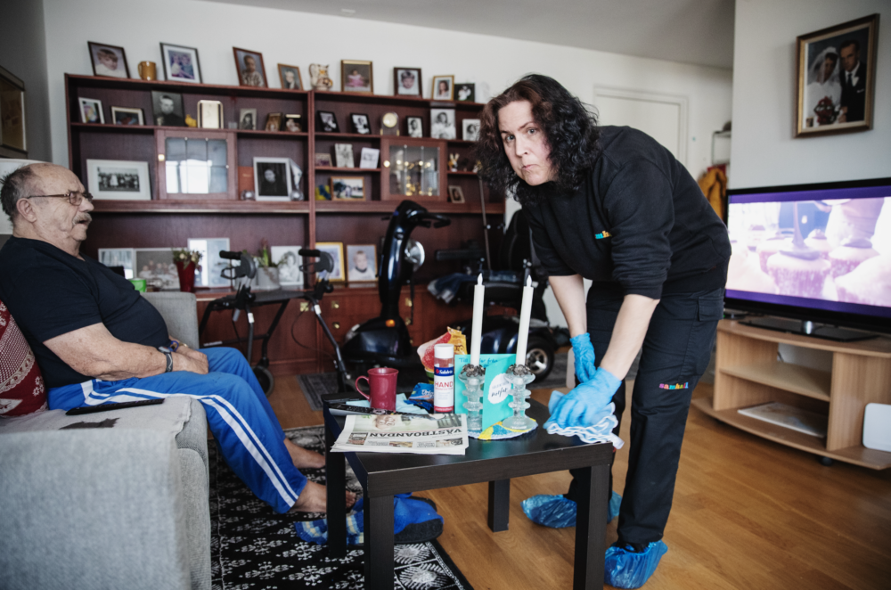 Monica Nilsson, i svart arbetsuniform, torkar ett vardagsrumsbord. I bakgrunden sitter en äldre man och tittar på tv.