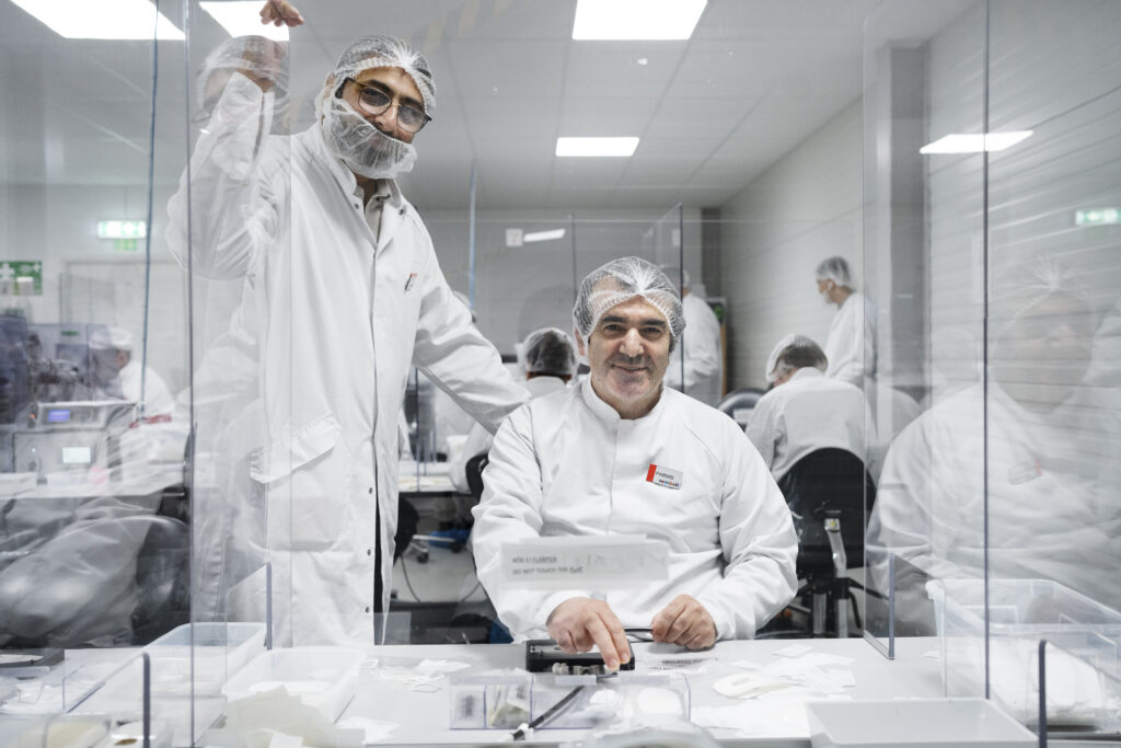 Bild på två personer i vita rockar och nätskydd på håret. De är omringdade av kollegor med samma arbetskläder. Rummet är vitt och arbetsytorna är avgränsade med glasrutor.