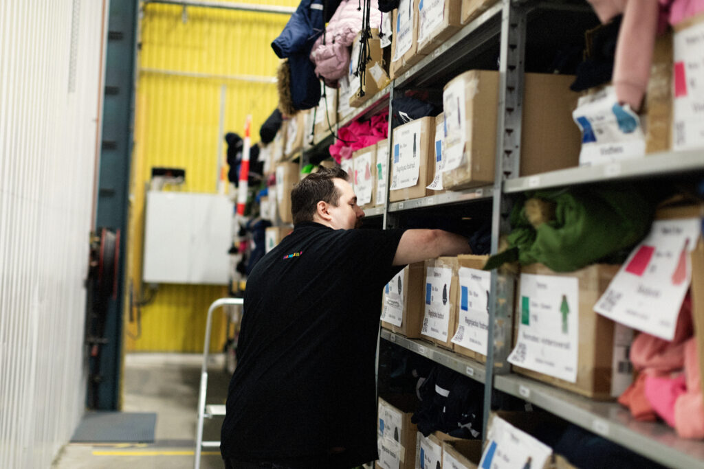 Jimmy står emd ryggen vänd mot karmen och rotar i en låd i ansiktshöjd på en lagerhylla. 