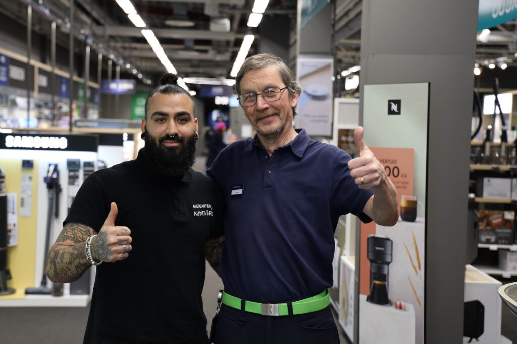 Bild på två män som står i en elgigantenbutik. De håller om varandra med en arm, den andra visar tummen upp. Båda tittar in i kameran och ler.