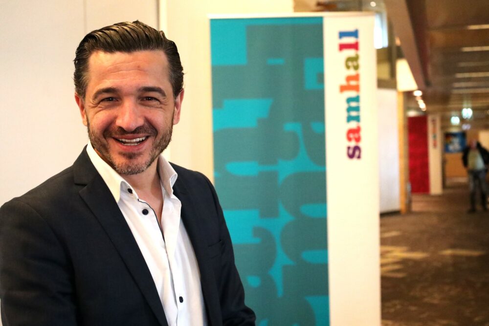 En man med kort mörkt hår i kostym framför en Samhall-rollup