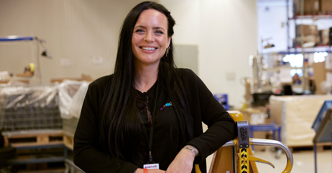 Porträtt av Jonna som har svart hår och svarta arbetskläder. Hon står lutad mot en gul palllyftare.