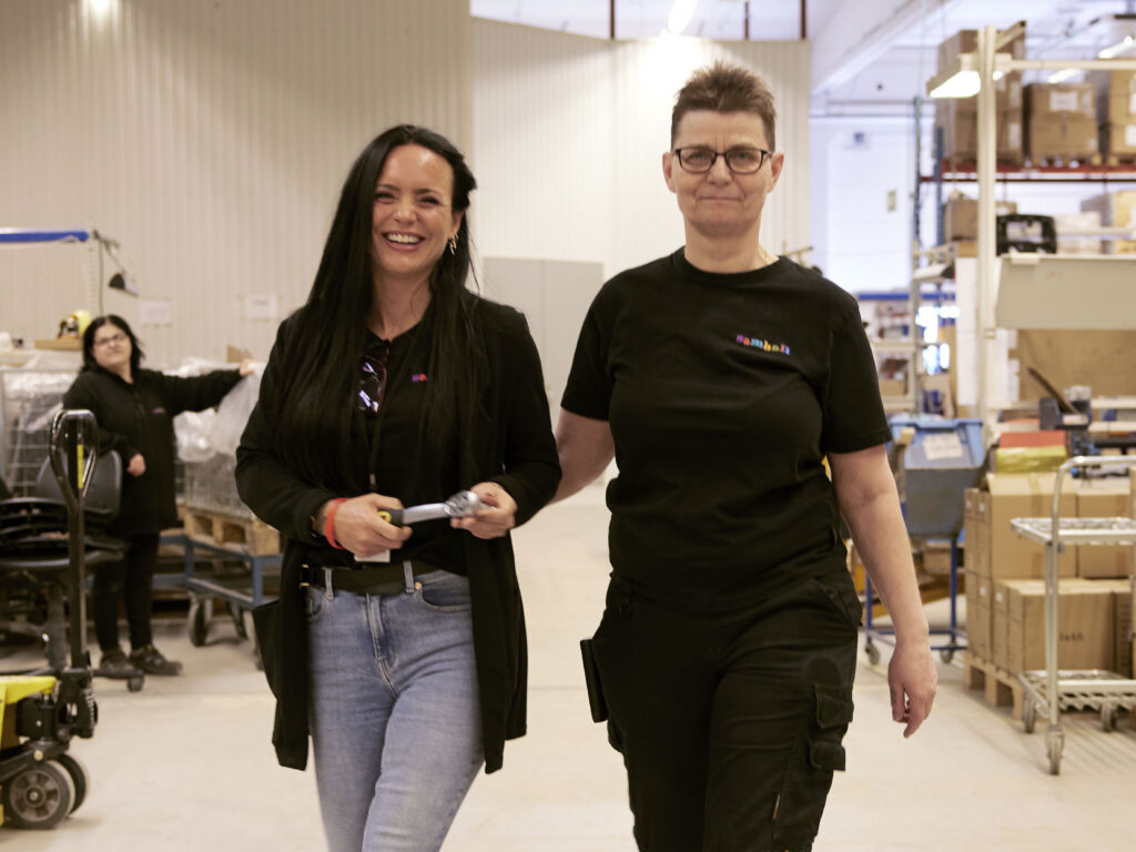 Bild på tre kvinnor i Samhallkläder. En i bakgrunden, två i förgrunden. De går i en lagerlokal.