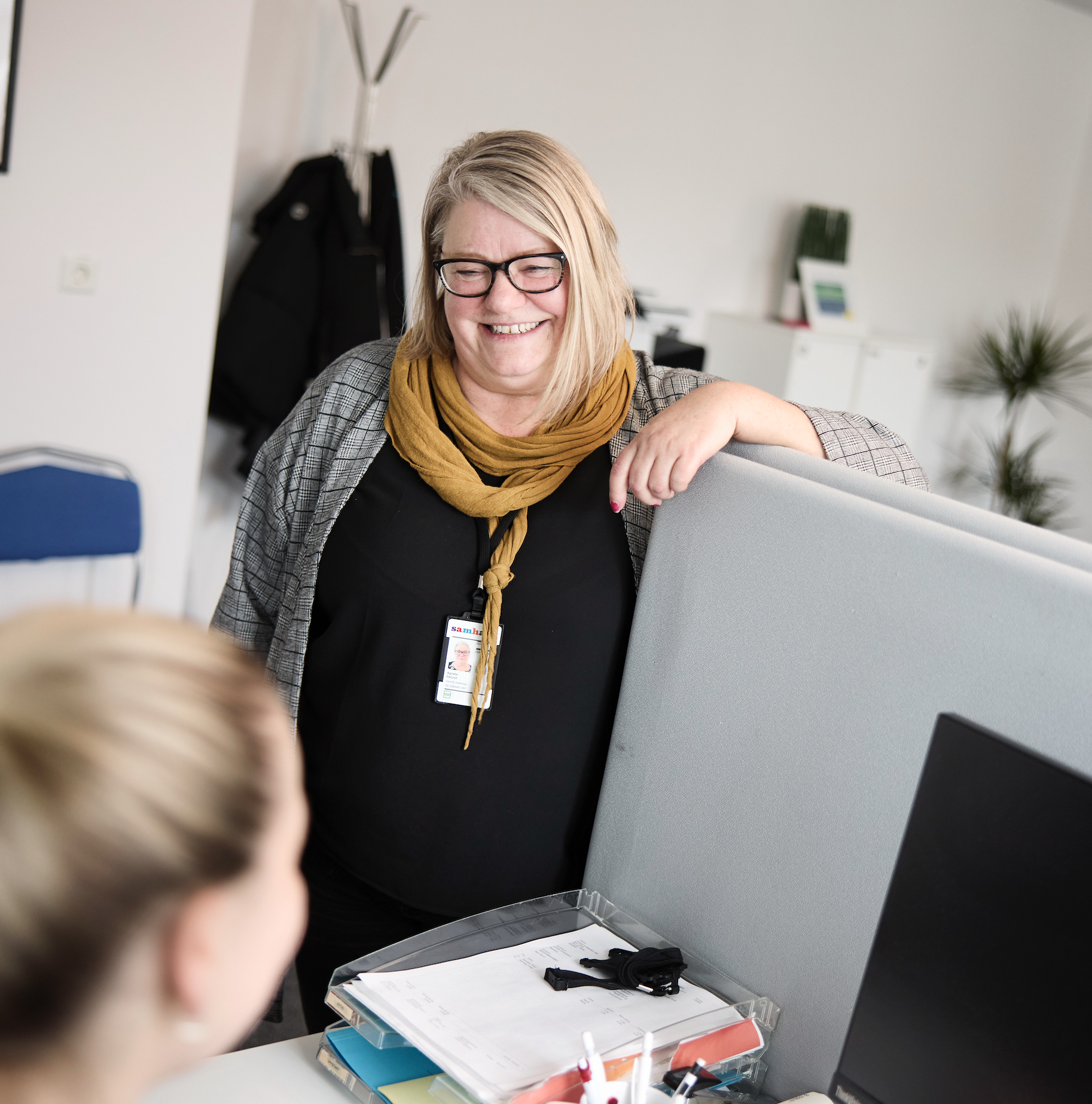 Bild på en kvinna som lutar ena armen på ett grått avskärmingsksydd vid en kontorsplats.