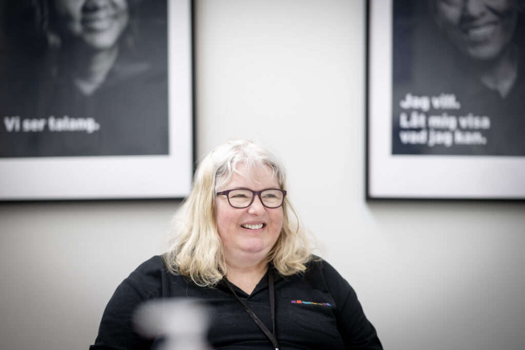 Kvinna med glasögon, ljust hår och svar Samhalltröja. Hon ler och tittar åt sidan. I bakgrunden syns två tavlor syddigt.