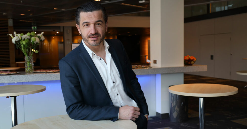 En man i kostym och svart hår står lutad mot ett småbord och tittar in i kameran.