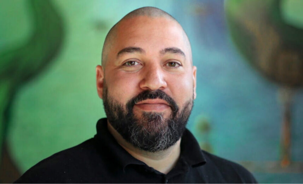 Porträtt på en man med rakat hår och mörkt skägg mot en grön bakgrund