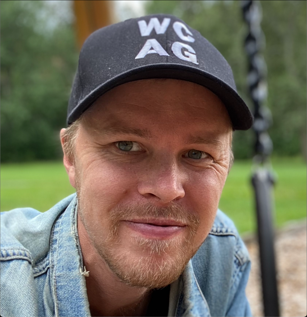 Bild på en man med jeansjacka och mörk keps. Han ler och tittar in i kameran.