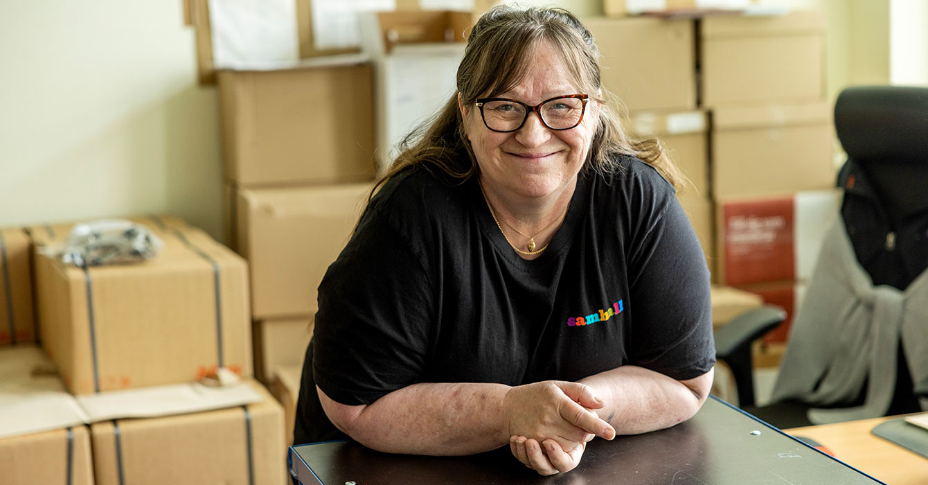 Porträtt på Inga-Lill som lutar sig mot ett arbetsbord. hon har långt hår och glasögon, bärandes svarta arbetskläder.