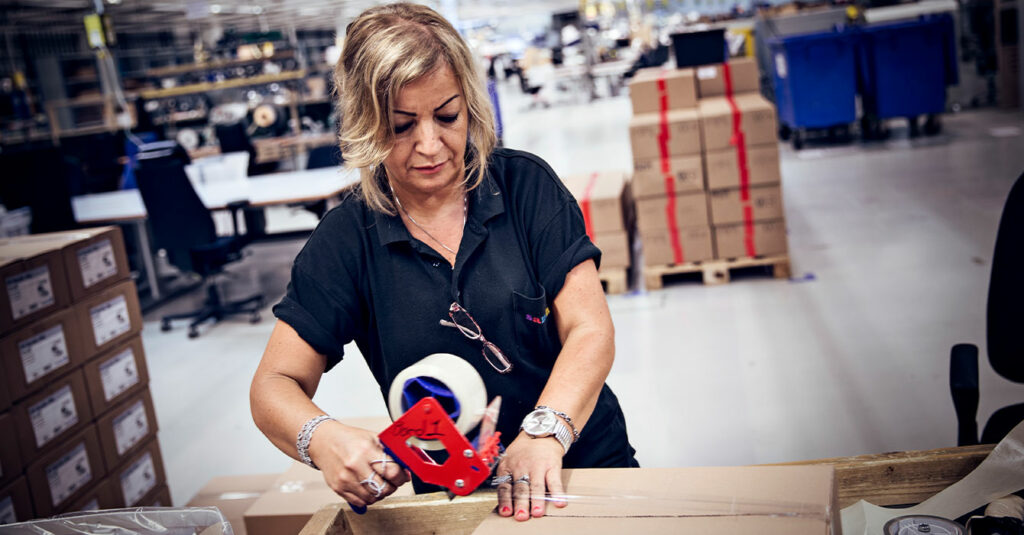 Bild på kvinna som står i ett lager och arbetar vid en arbetsbänk. hon tittar ner på bordet där det ligger ett paket som hon drar tejp över.