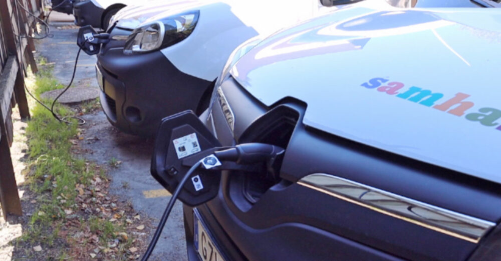 Närbild på en vit elbil som står och laddar med en sladd inkopplad. Man ser Samhall-loggan på motorhuven.