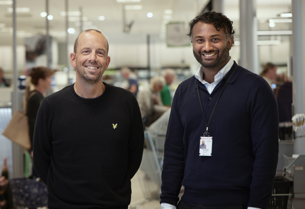 Två män står i en Sytembolaget-butik och tittar in i kameran. Båda ler.