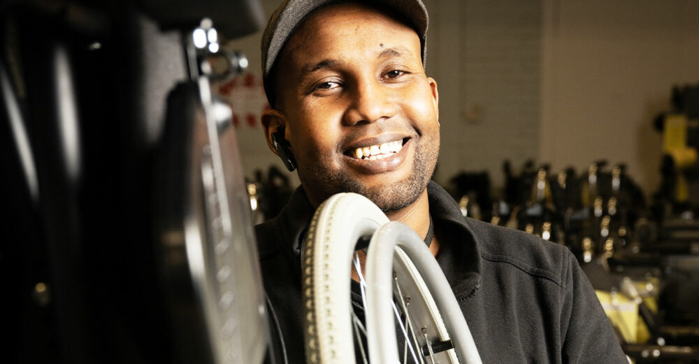 Bild på en person som står bakom ett rullstolsdäck. Han har mörka arbetskläder och keps.